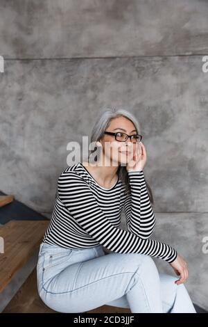 Foto di attraente donna asiatica con capelli grigi che indossa occhiali che guardano da parte mentre si siede su scala a casa Foto Stock