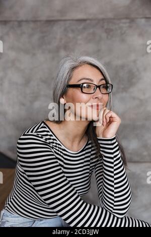 Foto di una donna asiatica con capelli grigi che indossa occhiali che guardano da parte mentre si siede su scala a casa Foto Stock