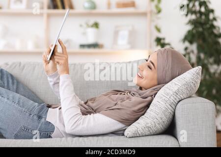 Passatempo di casa. Ragazza araba in hijab rilassante sul divano con tablet digitale Foto Stock