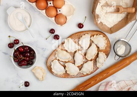 Preparare deliziosi gnocchi dolci con ciliegie. Dessert tradizionale russo e ucraino. Vareniki e ingredienti su fondo marmoreo -farina, Foto Stock