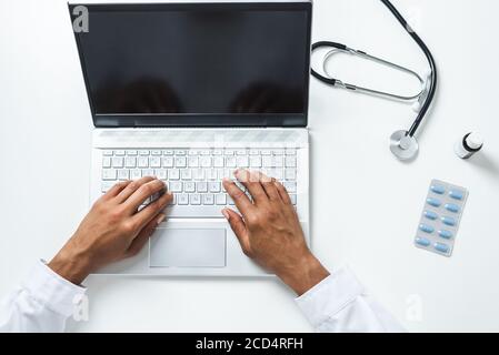 vista dall'alto delle mani di una dottoressa afro utilizzo di un computer portatile sulla parte superiore di una scrivania bianca con pillole e stetoscopio Foto Stock