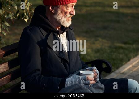 Vista laterale su senior bum male guardando disperatamente via vuota, in attesa di soldi, aiuto. Vestito con cappello rosso e cappotto caldo Foto Stock