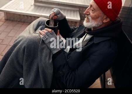 Mendicante uomo sedersi con vaso per soldi, indossando vecchi abiti caldi. Vagante con barba grigia, cappello rosso e guanti grigi Foto Stock