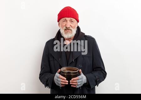 Il bum maturo in cappotto nero e il cappuccio rosso sulla testa che tiene la lattina di ferro per sollevare i soldi. Isolato su sfondo bianco Foto Stock