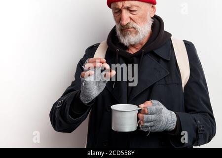 Uomo senza casa mature in cappotto nero e zaino bianco che tiene la tazza per raccogliere soldi e guardare la moneta. Isolato su sfondo bianco Foto Stock