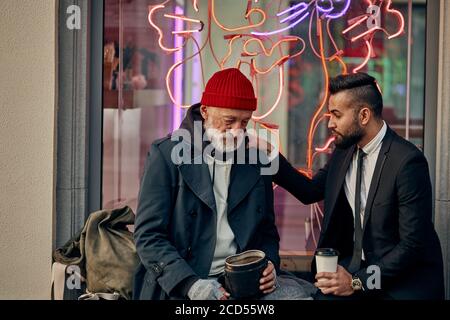 Uomo d'affari in tuxedo e senza casa in vestiti sporchi seduto e parlando in strada, guardare l'un l'altro. Povertà, aiuto Foto Stock