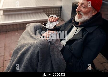 Mendicante uomo sedersi con vaso per soldi, indossando vecchi abiti caldi. Vagante con barba grigia, cappello rosso e guanti grigi Foto Stock