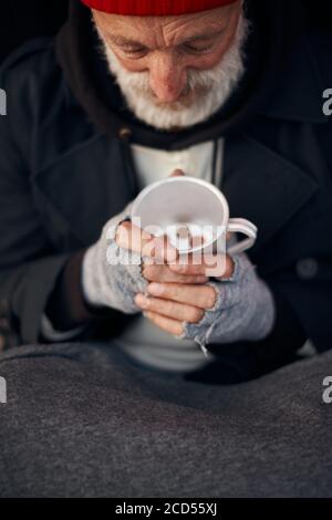 Mani di maschio senza casa maturo in guanti grigi che tengono la tazza con le monete. Povertà, fame, indifesi Foto Stock