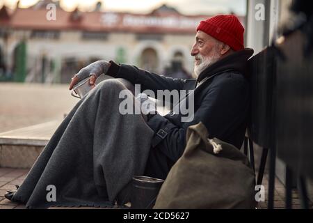 Vista laterale su uomo mendicante sulla strada che mendica, in attesa di aiuto da parte dei cittadini. L'uomo anziano senza dimora in abiti vecchi si siede sul pavimento in tempo freddo Foto Stock