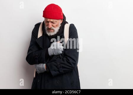 Bum maturo, senza casa vecchio uomo in cappotto e cappello rosso cercare di riscaldare isolato su sfondo bianco Foto Stock