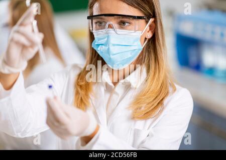 Donna medico che indossa maschera protettiva per il viso e googles di sicurezza siringa con ago da laboratorio e flaconcino di medicinale flacone di vaccino Foto Stock