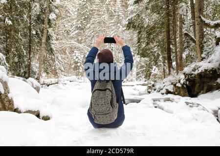 Ragazza con zaino seduto in una foresta di conifere innevata e scatta foto su uno smartphone. Giorno invernale. Vista posteriore Foto Stock