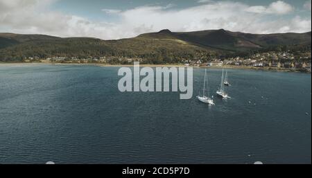 Golfo dell'oceano Atlantico, barche a vela zoom aereo sparato a Brodick Bay. Paesaggio scozzese della città portuale. Case, cottage, resort sulla riva del golfo contro terre verdi e vista sulle montagne Foto Stock