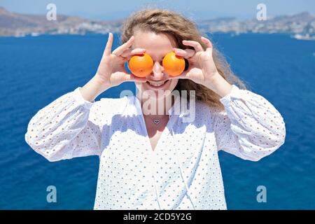 Bella donna in abito bianco che tiene oringes vicino ai suoi occhi Con vista mare sullo sfondo Foto Stock