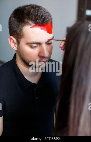 Ragazza artista dipinge su un viso mans. Pittura del viso sulla pelle. Ritratto di un giovane con vernice colorata su sfondo giallo. Trucco professionale F Foto Stock