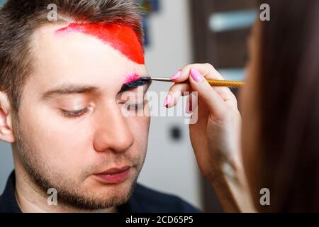 Ragazza artista dipinge su un viso mans. Pittura del viso sulla pelle. Ritratto di un giovane con vernice colorata su sfondo giallo. Trucco professionale F Foto Stock