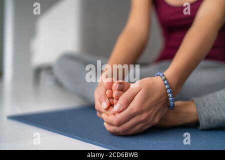 Yoga gamba allunga la donna a casa. Gambe a farfalla seduta allungano tenendo insieme le suole dei piedi. Closeup di mani che tengono i piedi nudi sul tappetino per esercizi Foto Stock