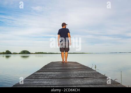 Uomo in pantaloni corto carico in piedi su molo di legno e osservando la superficie del lago, Varazdin lago, Croazia Foto Stock