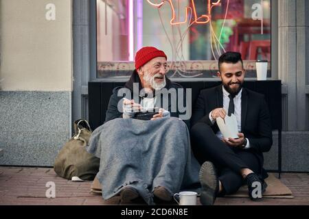 Sorridendo ricchi e poveri uomini insieme seduti sulla strada e mangiare mentre parlano. Felice nonostante la disuguaglianza sociale Foto Stock