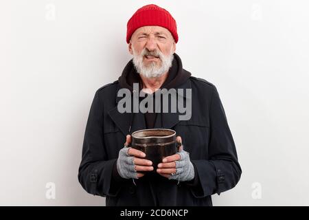 Il bum maturo in cappotto nero e il cappuccio rosso sulla testa che tiene la lattina di ferro per sollevare i soldi. Isolato su sfondo bianco Foto Stock