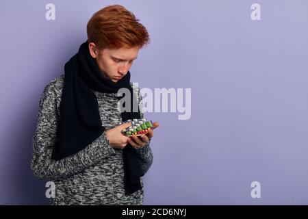 L'uomo arrossito dai capelli rossi si sente male, , ha sintomi di freddo, soffre di febbre, sceglie pillole, malvagità. Copia spazio. Primo piano ritratto. Isolato bac blu Foto Stock
