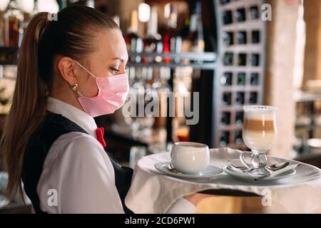 Una cameriera femminile di aspetto europeo in una maschera medica serve caffè latte. Foto Stock