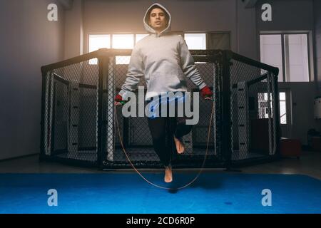 Sportivo in grigio felpa con cappuccio esercizi con corda di salto vicino all'anello di pugilato, riscaldamento prima di combattere Foto Stock