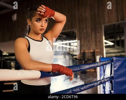 Boxer donna stanca dopo un intenso allenamento in anello, stare appoggiandosi sulla recinzione, efficace allenamento in palestra. Foto Stock