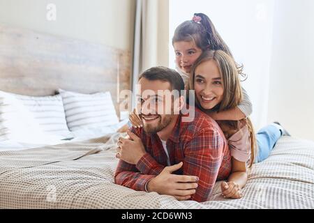 Positivo ottimista uomo sdraiato sul letto in una comoda camera d'albergo, tenendo le mani incrociate, guardandosi via, moglie e bambino si riposano felicemente sulla schiena, Foto Stock