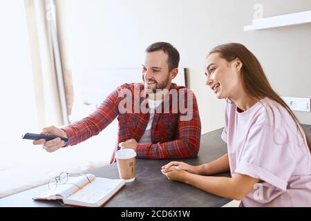 Piacevole marito e moglie molto gentili seduti al tavolo della camera d'albergo, guardando la tv, chiacchierando, scegliendo un buon film, girando in interni, ritratto Foto Stock