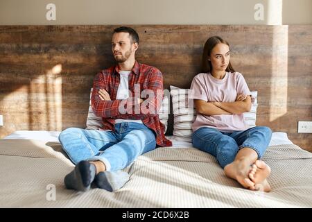 L'uomo insoddisfatto non rasato indossa una maglietta rossa corta, bianca, jeans blu e calzini, seduto sul letto che esprime irritazione dopo il litigio con la giovane donna si Foto Stock