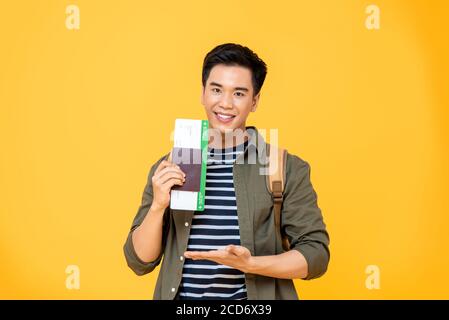 Ritratto di sorridente giovane asiatico zaino in spalla turista uomo mostrando suo passaporto con carta d'imbarco in gesto palmo aperto isolato studio backgrou giallo Foto Stock