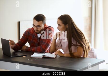 Affascinante donna attraente si siede vicino da un bell'uomo a tavola, ascoltarlo con attenzione, esprimendo interesse, tenendo il mento, guardando lo schermo con Foto Stock