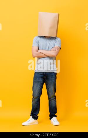 Concept ritratto a lunghezza intera dell'uomo anonimo con testa coperta con sacchetto di carta che fa le braccia incrociate gesto in studio giallo sfondo Foto Stock