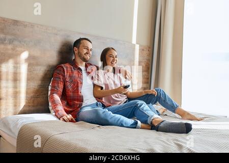 Una coppia giocosa che sorride guardando un programma televisivo divertente, sorridendo ampiamente, uomo non rasato che mette mano sulle spalle della sua giovane moglie, ritratto, conca di felicità Foto Stock