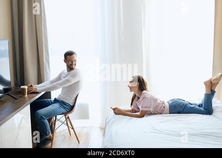 Felice coppia con il computer portatile trascorrere il tempo insieme a casa, uomo piacevole si siede sulla sedia, si gira la testa alla giovane moglie, guarda via positivamente, parlando di finta Foto Stock