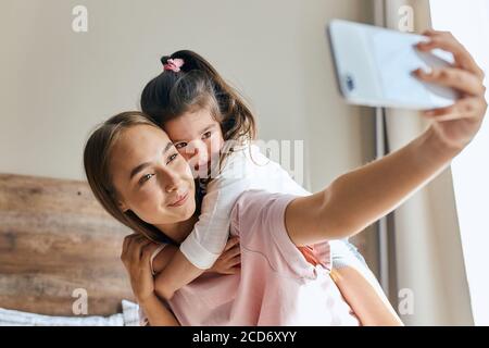 Affascinante giovane donna prende selfie utilizzando il telefono cellulare, carino allegro bambino hugs mamma, esprimendo la felicità, guardando da parte, si concentra in dispositivo, primo piano, pos Foto Stock