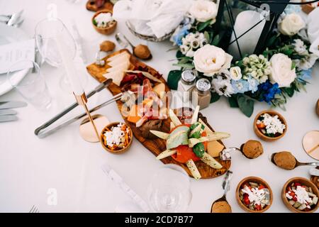 Tagliere con prelibatezze sul tavolo con ciotole, shaker di sale, shaker di pepe e una grande candela in un bouquet di fiori. Foto Stock