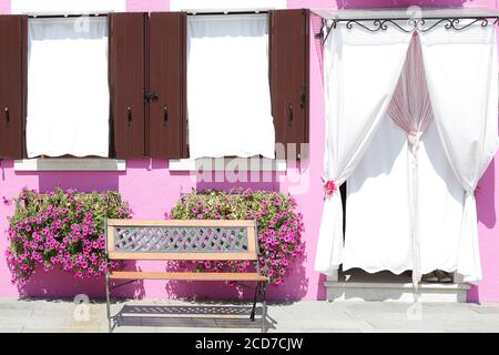 Parete dipinta rosa con due finestre con persiane in legno e una porta coperta con una tenda sul davanzale, una in vaso con fiori, all'isola di B. Foto Stock