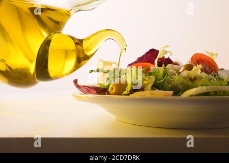 Versare l'olio d'oliva dal distributore dell'olio sull'insalata preparata piastra bianca e sfondo isolato Foto Stock