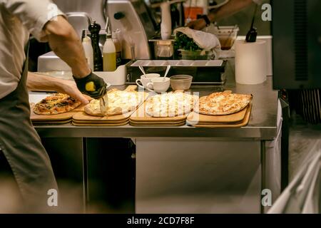 Cucina pizzeria. Cuoco cuoco in grembiule e guanti tagliano la pizza appena sfornata con coltello a rullo. Foto Stock