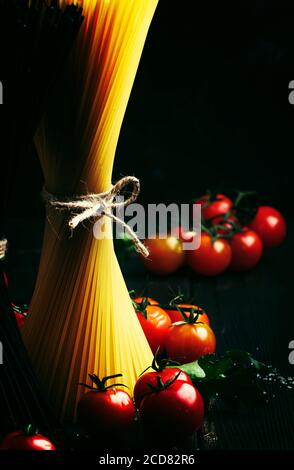 Spaghetti di pasta nero e giallo, legati con corda, si trovano verticalmente su un tavolo da cucina scuro con ingridenti per la cottura, bassa vita morta, selettivi f Foto Stock