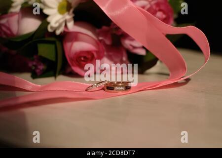 Anelli di nozze su un nastro di seta rosa su uno sfondo rosa bouquet. Primo piano Foto Stock