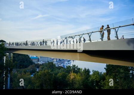 Immagine fuori fuoco, un ponte moderno con la gente sopra un fiume recintato da una recinzione di vetro contro un cielo blu chiaro. Foto Stock