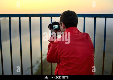 Un uomo su un ponte sul fiume che fotografa un'alba su una nebbia mattina presto. Foto Stock