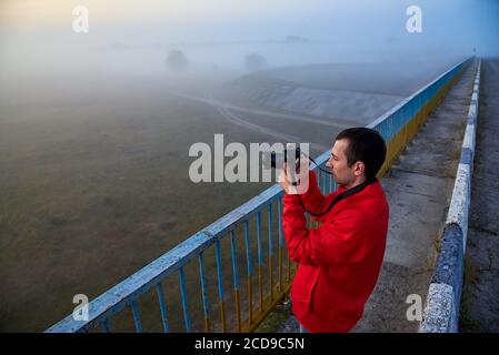 Un uomo su un ponte sul fiume che fotografa un'alba su una nebbia mattina presto. Foto Stock