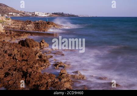 Il movimento delle onde catturate nella luce del mattino presto con un pescatore solista sulla costa rocciosa di Stalis, Creta, Grecia. Foto Stock