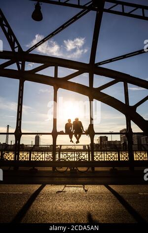 Persone retroilluminate non identificabili che godono del sole serale seduto su un ponte di acciaio sopra i binari ferroviari chiamato Haccurbrücke. Monaco, Germania Foto Stock