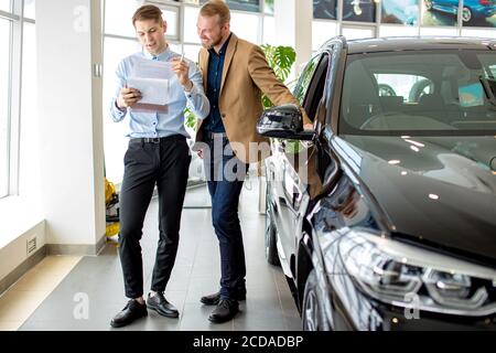 giovane venditore di automobili caucasiche mostrano la migliore variante al cliente in concessionaria. bell'uomo d'affari è venuto per ottenere nuovo auto. consulente aiutarlo con ch Foto Stock