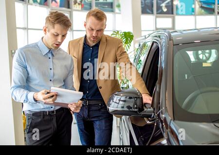 bel consulente caucasico affabile parlare di caratteristiche e vantaggi di auto in concessionaria, giovane uomo è venuto a ottenere nuova automobile, scegliere e ascoltare Foto Stock